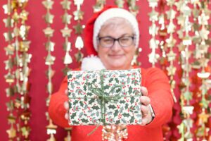 A red and gold Christmas background. Fiona is stood in a Christmas jumper and hat, holding out a wrapped Christmas present.