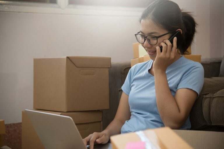 A young professional making a call while working on a laptop amidst moving boxes, representing the journey of building an online business with Darren and Mike’s mentorship and support.