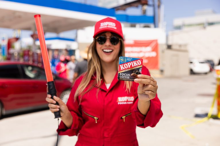A woman in a red uniform and cap smiles while holding a Kopiko Focus Coffee packet and a red baton. A lively gas station scene is in the background.