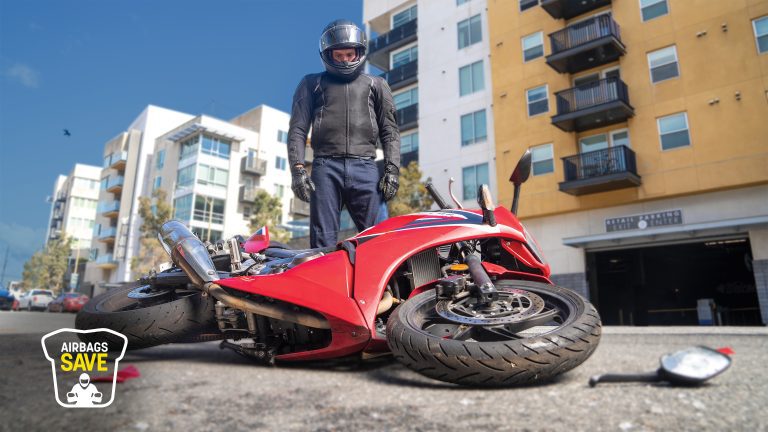 A motorcyclist in a helmet and wearable airbag stands beside a crashed red motorcycle on a city street. Nearby, a broken mirror lies on the ground, emphasizing the importance of motorcycle safety.